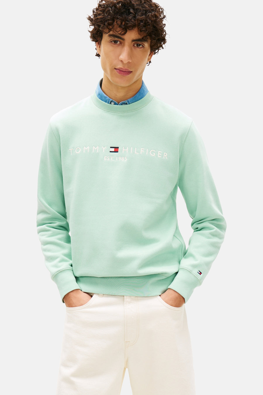 Sweat-shirt vert à col rond de TOMMY HILFIGER, avec logo brodé, porté sur une chemise en denim et un pantalon blanc.
