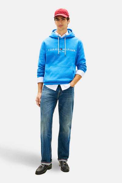 Sweat-shirt bleu à capuche de Tommy Hilfiger avec logo imprimé, porté sur une chemise rayée et un jean, avec un chapeau rouge.