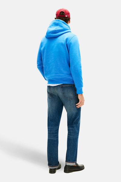 Sweat à capuche bleu de TOMMY HILFIGER, porté avec un jean et des mocassins noirs, coiffé d'une casquette rouge.