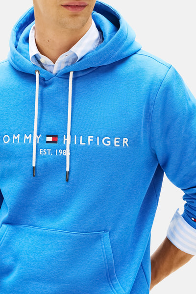 Blauwe Tommy Hilfiger hoodie, met witte trekkoord en geborduurd logo op de voorkant, gedragen over een gestreept overhemd.
