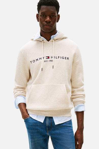 Sweat écru à capuche de Tommy Hilfiger, avec cordon, porté sur une chemise rayée bleu et un jean.