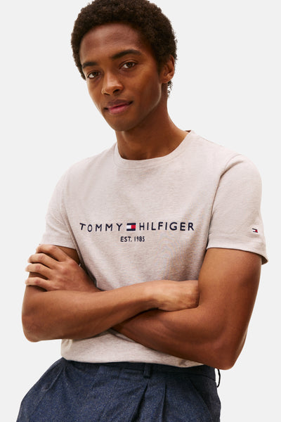 T-shirt beige à manches courtes de TOMMY HILFIGER, avec logo sur le devant, porté avec un pantalon bleu marine.

