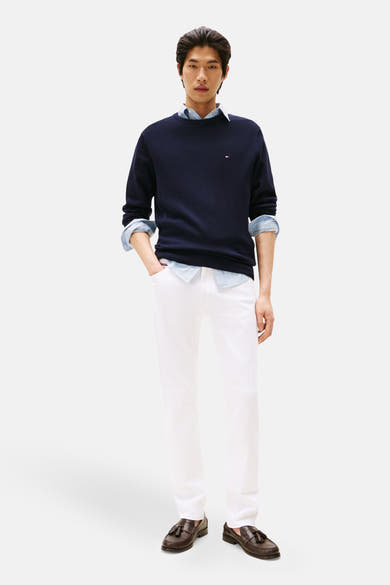 Pull à col rond bleu de Tommy Hilfiger, porté avec une chemise claire et un pantalon blanc.