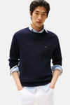 Pull à col rond bleu de Tommy Hilfiger, porté sur une chemise boutonnée à rayures, avec un pantalon blanc.