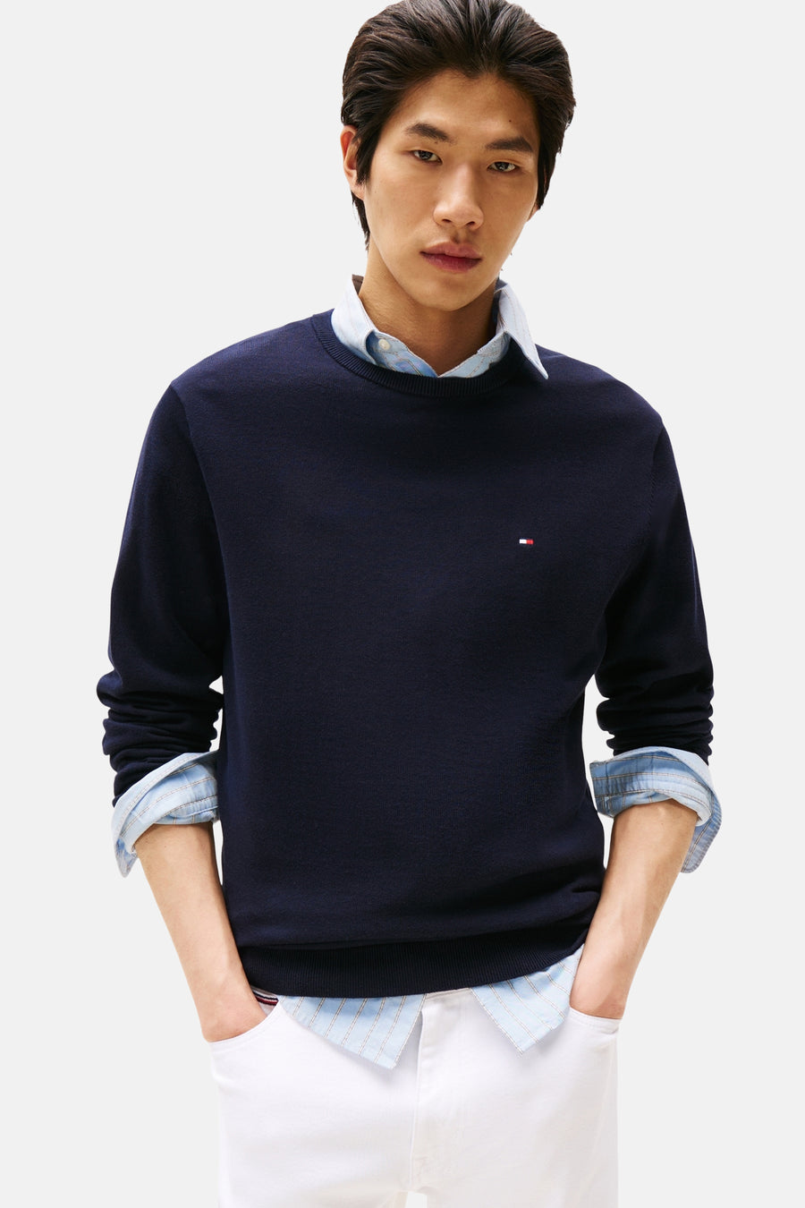 Pull à col rond bleu de Tommy Hilfiger, porté sur une chemise boutonnée à rayures, avec un pantalon blanc.