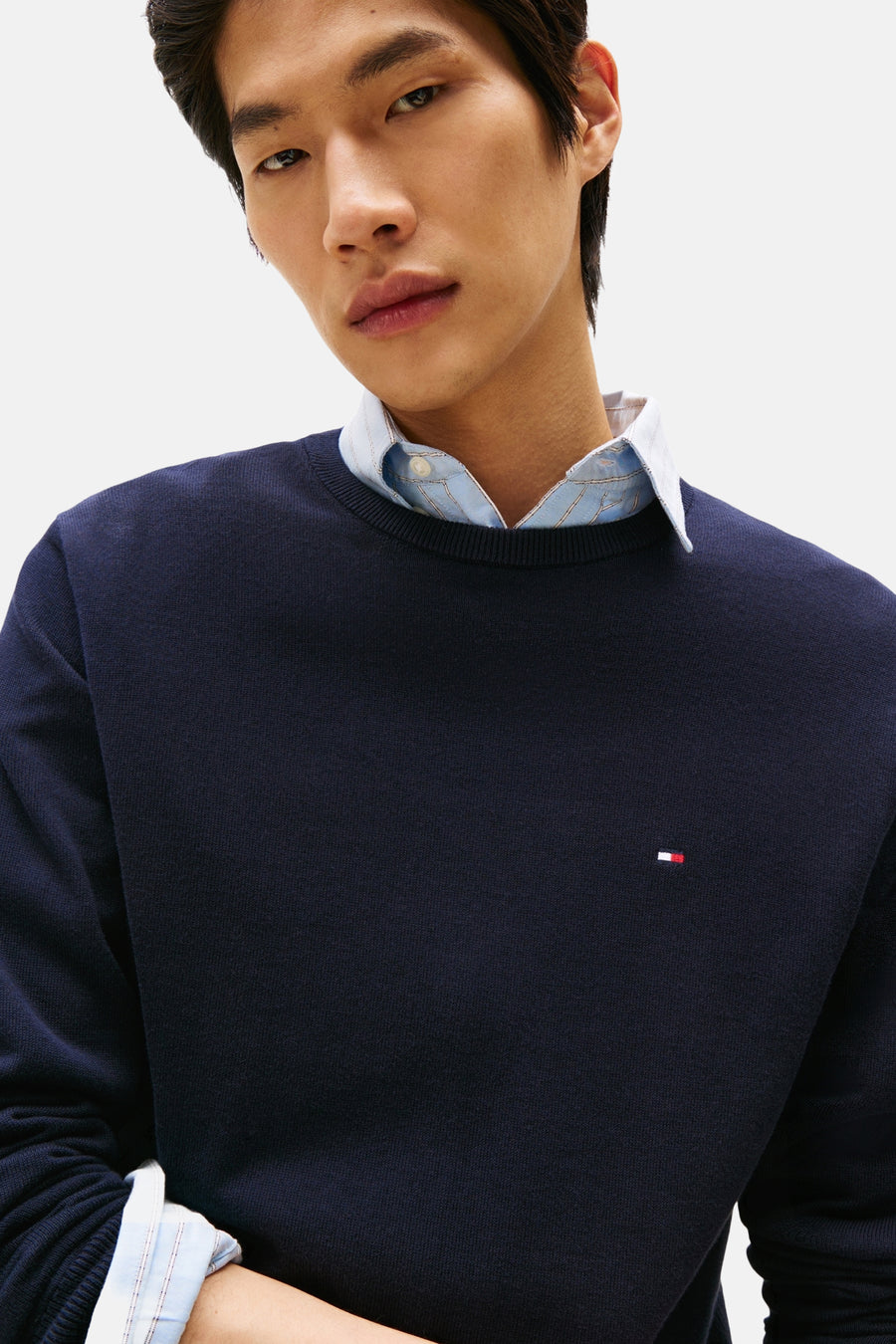 Pull à col rond bleu de Tommy Hilfiger, porté sur une chemise à rayures bleu clair et blanches.