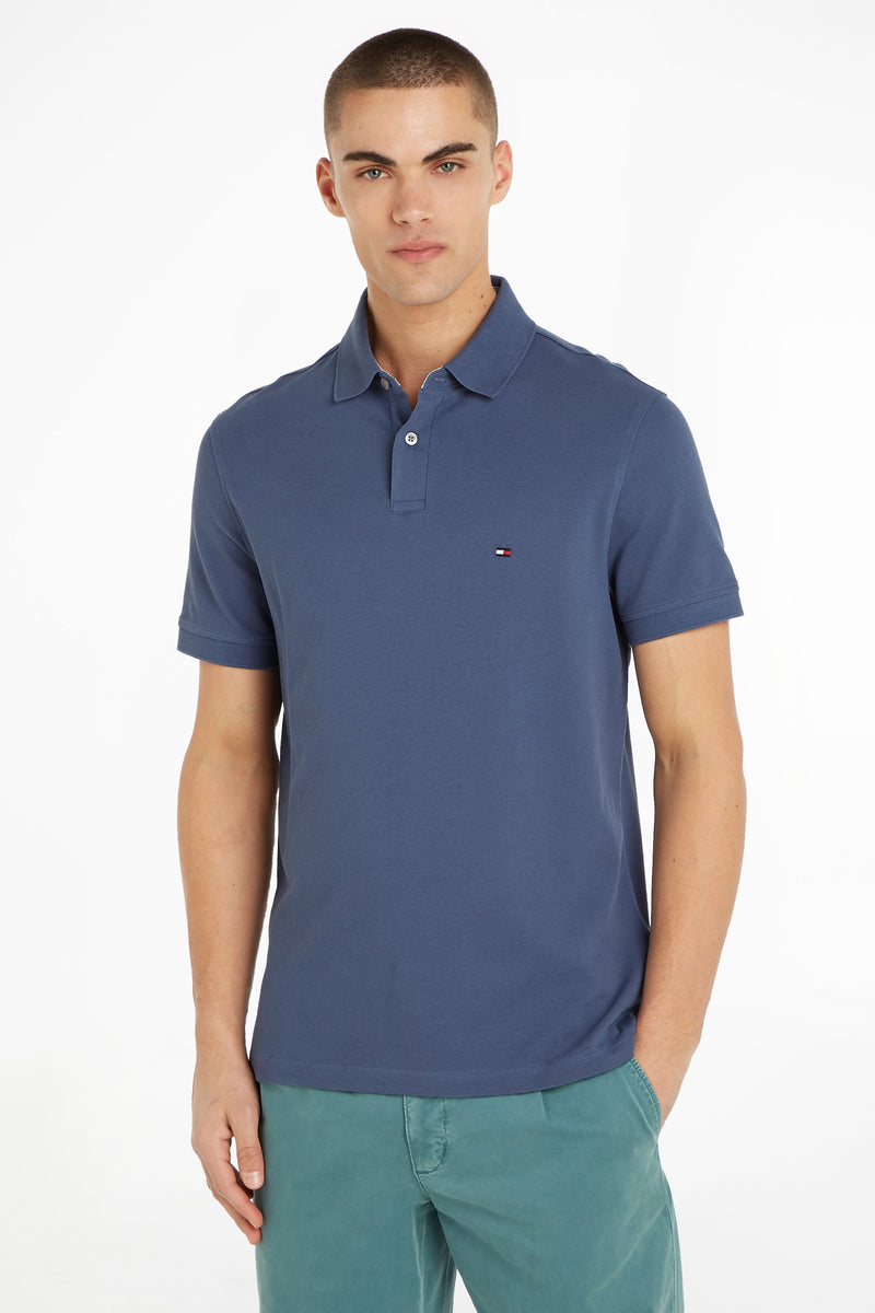 Blue short-sleeve polo shirt by Tommy Hilfiger, featuring ribbed texture and small flag logo on chest, worn with teal pants in front view.