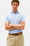 Polo à manches courtes bleu par Tommy Hilfiger, porté avec un pantalon beige et une ceinture marron, bras croisés.
