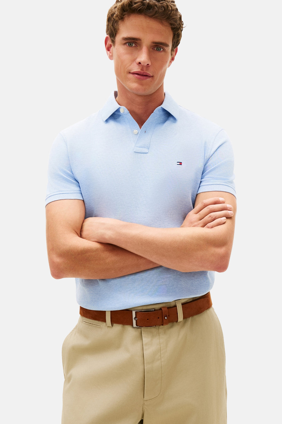 Polo à manches courtes bleu par Tommy Hilfiger, porté avec un pantalon beige et une ceinture marron, bras croisés.