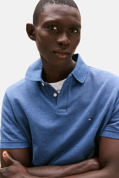 Polo bleu à manches courtes de TOMMY HILFIGER, sur un mannequin au teint foncé, bras croisés.
