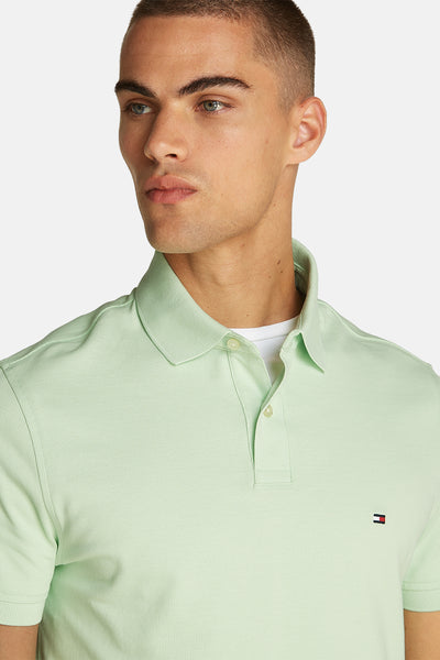 Polo vert à manches courtes de Tommy Hilfiger avec logo sur la poitrine, porté sur un t-shirt blanc.