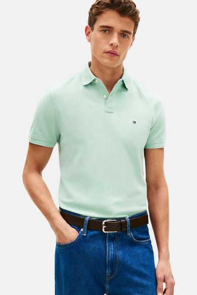 Polo à manches courtes vert de TOMMY HILFIGER avec logo, porté avec un jean bleu et une ceinture marron.
