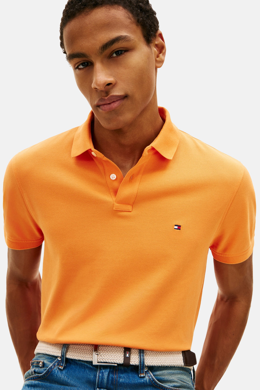 Polo à manches courtes orange de Tommy Hilfiger, porté avec un jean et une ceinture tressée.