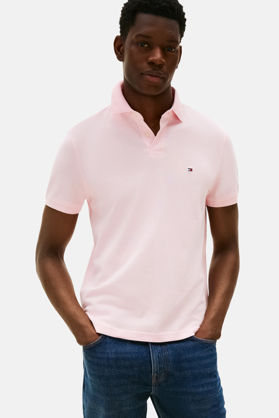 Polo rose à manches courtes de Tommy Hilfiger, porté avec un jean bleu, les mains dans les poches.

