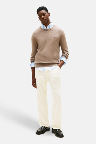 Beige ronde hals trui van TOMMY HILFIGER, gedragen over een lichtblauw gestreept overhemd, met gebroken witte broek en zwarte loafers.