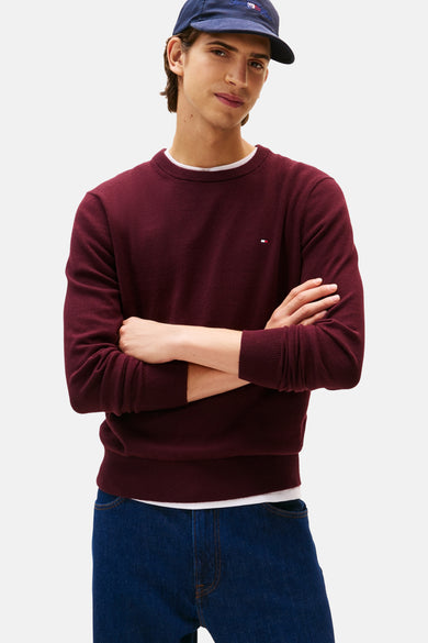 Bordeaux ronde hals trui van TOMMY HILFIGER, gedragen over een wit T-shirt en een blauwe jeans, met een marineblauwe pet.