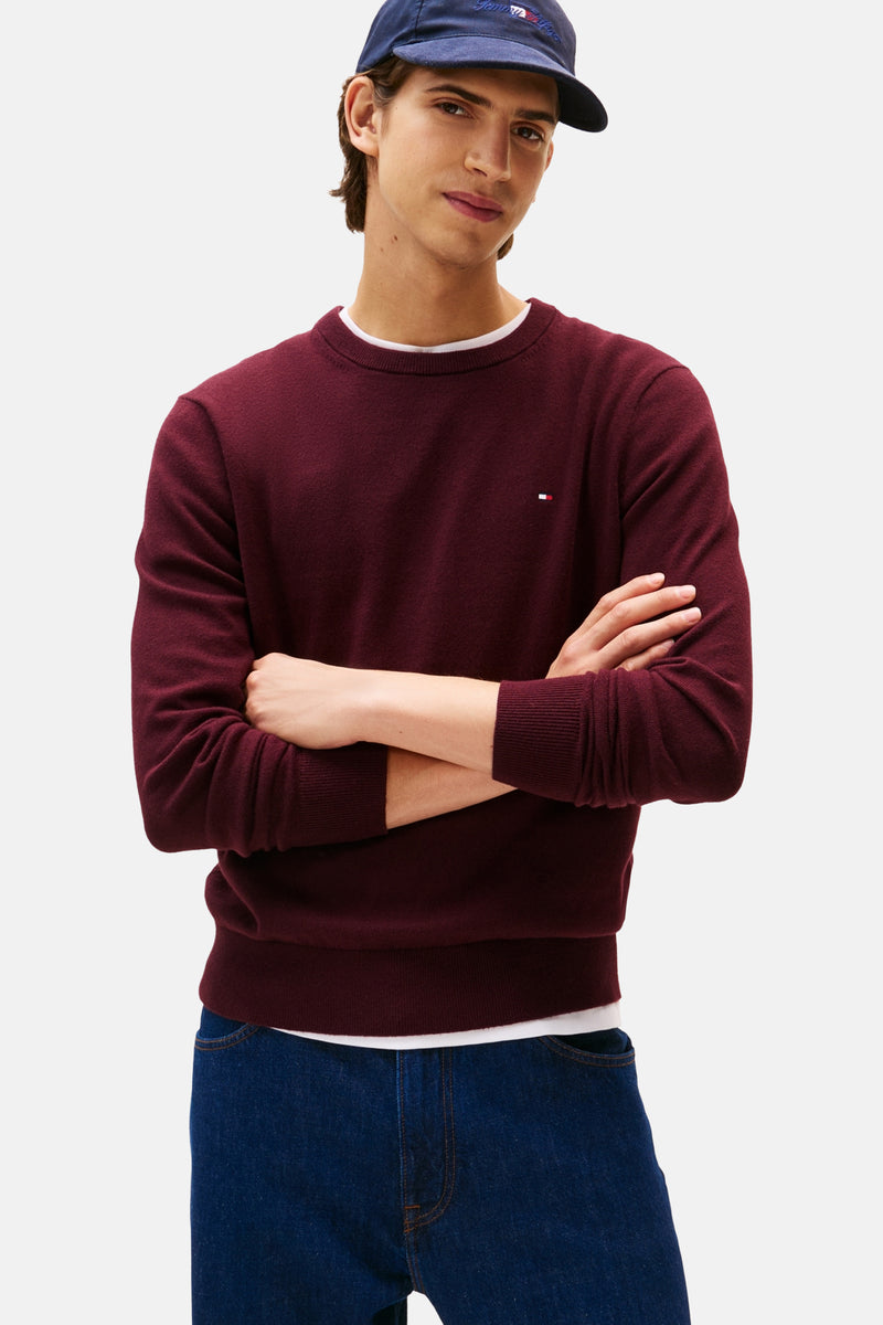 Bordeaux ronde hals trui van TOMMY HILFIGER, gedragen over een wit T-shirt en een blauwe jeans, met een marineblauwe pet.