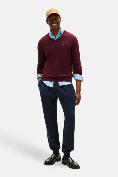 Bordeaux V-hals trui van TOMMY HILFIGER, gedragen over een lichtblauw overhemd, met een marineblauwe joggingbroek en zwarte loafers.