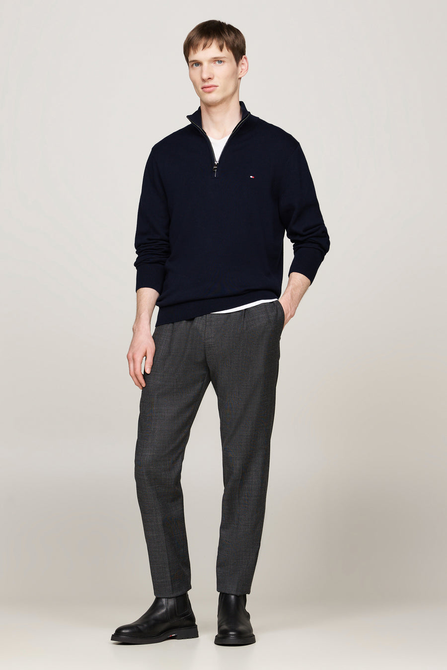 Pull bleu à col zippé de Tommy Hilfiger porté avec un pantalon gris à motifs et des bottines noires.