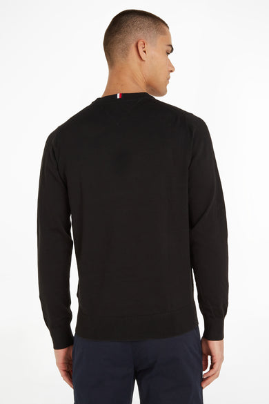 Pull col rond noir de TOMMY HILFIGER, avec détails tricolores à l'arrière du cou, porté avec un pantalon bleu marine.
