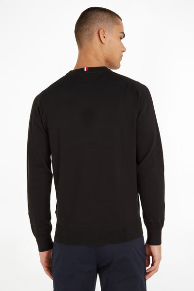 Pull col rond noir de TOMMY HILFIGER, avec détails tricolores à l'arrière du cou, porté avec un pantalon bleu marine.
