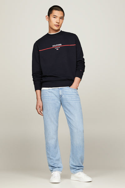 Sweat-shirt bleu marine de Tommy Hilfiger, avec un logo rouge et blanc sur la poitrine, porté avec un jean clair et des baskets blanches.
