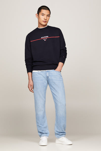 Blauwe trui met ronde hals van Tommy Hilfiger, met een rode bies en het logo op de voorkant, gedragen met een spijkerbroek en witte sneakers.