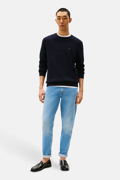 Pull à col rond bleu de TOMMY HILFIGER, porté sur un t-shirt blanc, avec un jean bleu et des mocassins noirs.
