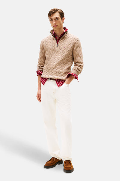 Pull col montant beige en maille torsadée de Tommy Hilfiger, porté sur une chemise rayée rouge et blanc, avec un pantalon blanc.