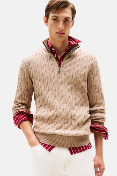 Pull à col roulé beige de TOMMY HILFIGER, avec motif torsadé, porté sur une chemise rouge rayée, pantalon blanc.
