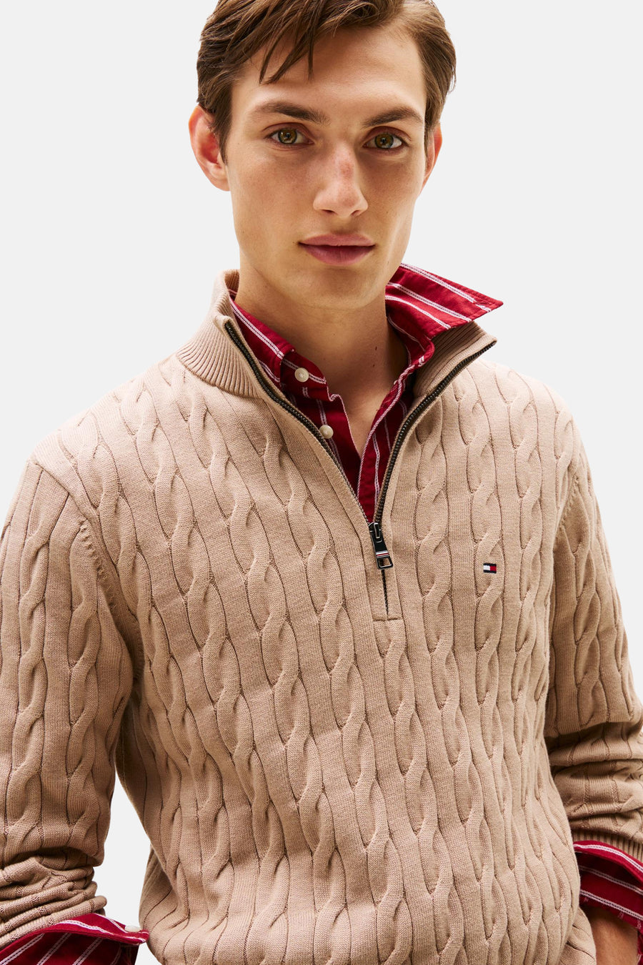Beige coltrui van TOMMY HILFIGER met kabelpatroon en ritssluiting, gedragen over een gestreept rood overhemd.