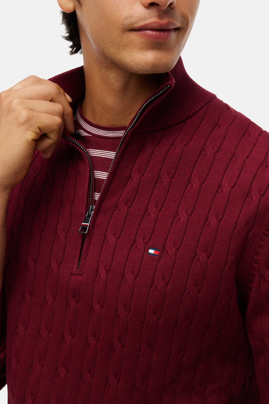 Burgundy cable-knit turtleneck sweater by Tommy Hilfiger, featuring front zipper and small flag logo on chest, worn over striped tee, close-up front view