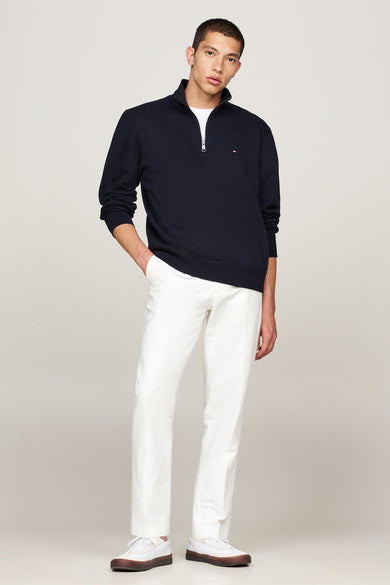 Blauwe sweater met opstaande kraag van TOMMY HILFIGER, met ritssluiting en discreet logo, gedragen over een wit T-shirt en een witte broek.