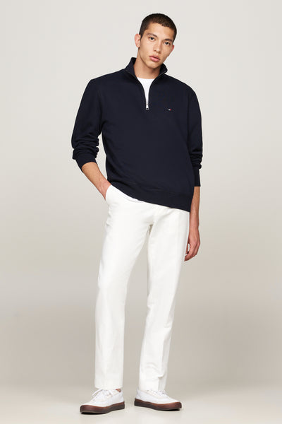 Blauwe sweater met opstaande kraag van TOMMY HILFIGER, met ritssluiting en discreet logo, gedragen over een wit T-shirt en een witte broek.