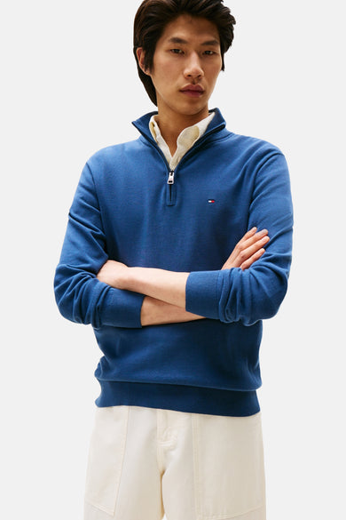 Pull bleu à col roulé de Tommy Hilfiger, porté sur une chemise blanche, les bras croisés.