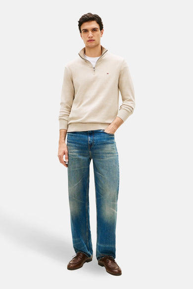 Pull à col roulé beige de TOMMY HILFIGER, porté avec un t-shirt blanc et un jean bleu délavé, avec des chaussures marron.