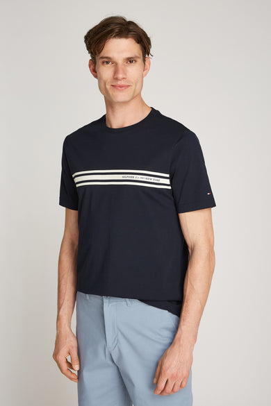 T-shirt à manches courtes bleu marine de Tommy Hilfiger avec rayures blanches horizontales sur le devant, porté avec un pantalon bleu.