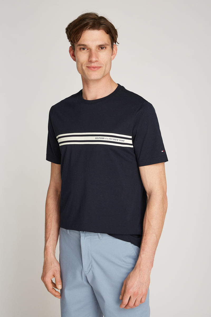 T-shirt à manches courtes bleu marine de Tommy Hilfiger avec rayures blanches horizontales sur le devant, porté avec un pantalon bleu.