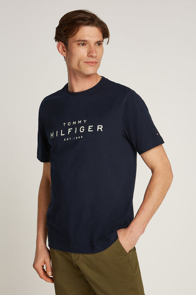 T-shirt bleu à manches courtes de TOMMY HILFIGER, avec logo blanc sur le devant, porté avec un pantalon kaki.