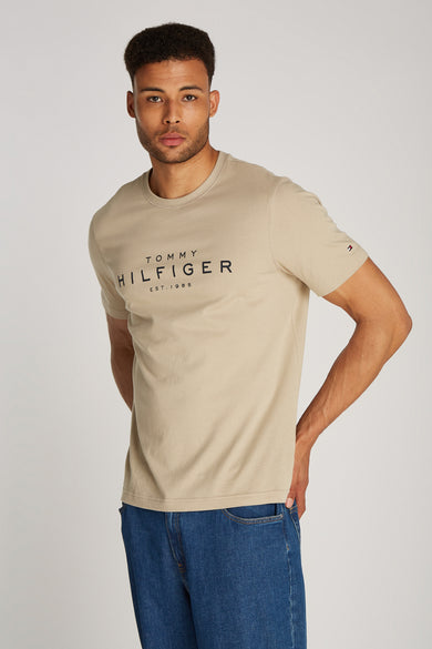 T-shirt beige à manches courtes de TOMMY HILFIGER, avec logo imprimé sur le devant, porté avec un jean bleu.