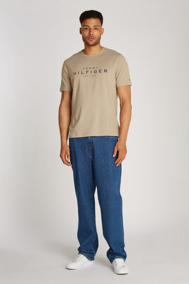 T-shirt à manches courtes beige de TOMMY HILFIGER, porté avec un jean bleu et des baskets blanches.
