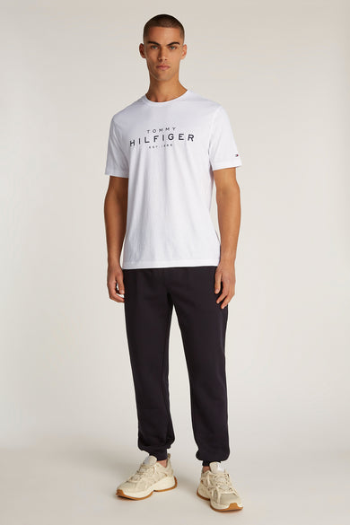T-shirt à manches courtes blanc de TOMMY HILFIGER, avec logo imprimé sur le devant, porté avec un jogging noir et des baskets.