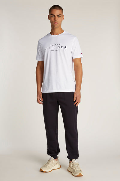 T-shirt à manches courtes blanc de TOMMY HILFIGER, avec logo imprimé sur le devant, porté avec un jogging noir et des baskets.