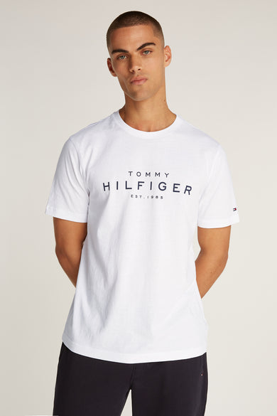 T-shirt blanc à manches courtes de TOMMY HILFIGER, avec logo centré sur le devant, porté avec un short noir uni.