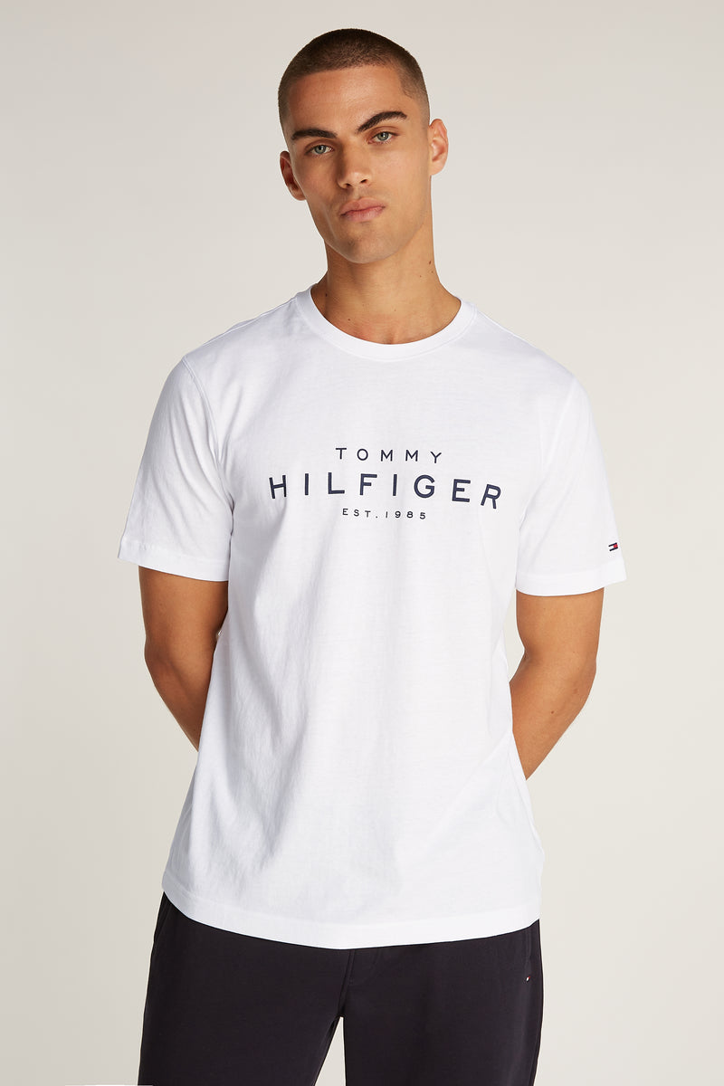 T-shirt blanc à manches courtes de TOMMY HILFIGER, avec logo centré sur le devant, porté avec un short noir uni.