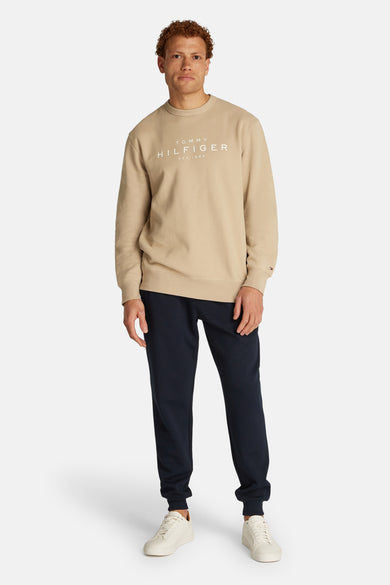 Sweat-shirt beige de TOMMY HILFIGER, avec logo blanc sur le devant, porté avec un pantalon de survêtement bleu marine et des baskets blanches.
