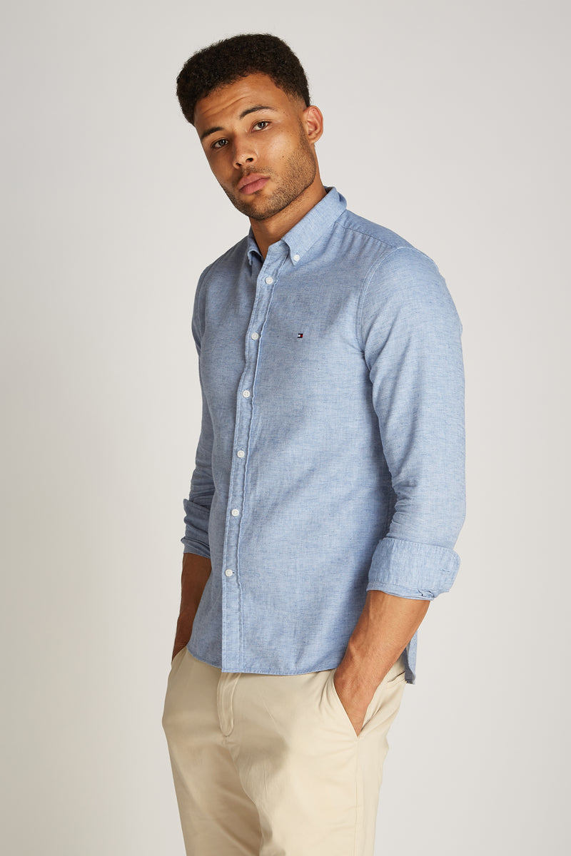 Chemise à manches longues bleu de TOMMY HILFIGER, portée avec un pantalon beige, avec les mains dans les poches.