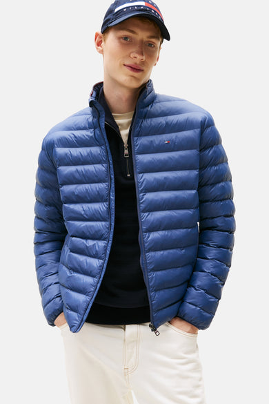 Veste courte bleue de Tommy Hilfiger avec fermeture éclair et logo sur la poitrine, portée sur un pull et un pantalon blanc.
