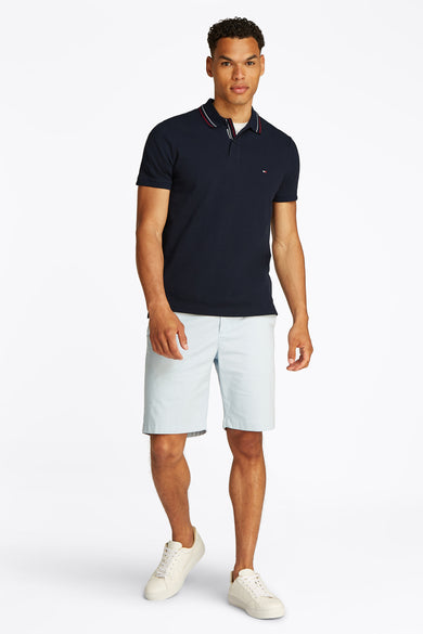 Marineblaues Poloshirt mit kurzen Ärmeln von TOMMY HILFIGER, getragen mit einer hellblauen Shorts und weißen Turnschuhen.