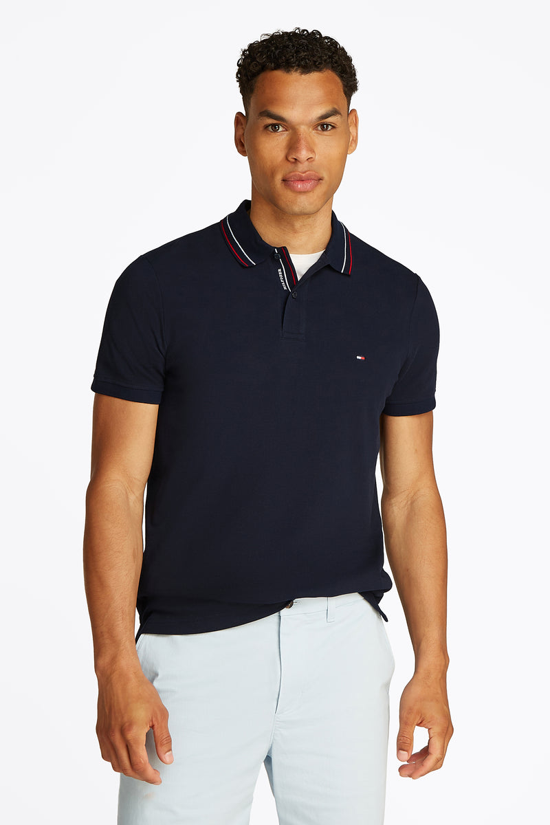 Donkerblauwe polo met korte mouwen van Tommy Hilfiger, rood-wit gestreepte kraag, gedragen met een lichtblauwe broek.